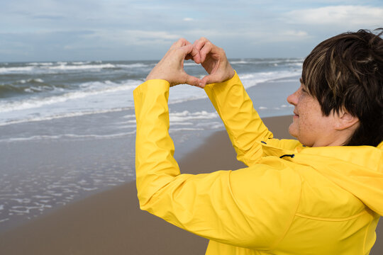 Smiling woman with freckles showing heart sign on sea. Yellow raincoat clothe for sea walk. Love sign