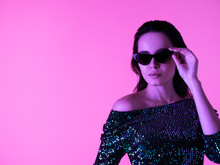 Young beautiful confident brunette female in trendy evening shiny green reflecting dress. Sexy carefree woman isolated on pink neon light in studio. Fashionable model in sunglasses. High fashion