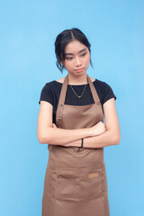 A young asian female barista feeling down and sad. Wearing a brown apron and black shirt and against a light blue background.