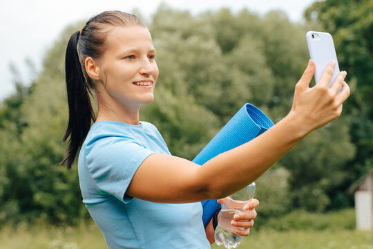 Happy millennial woman runner in sportswear take selfie on mobile phone in green forest park.Candid girl at workout fitness exercise hold yoga mat, water bottle.Girl in trendy pink blue fitness wear