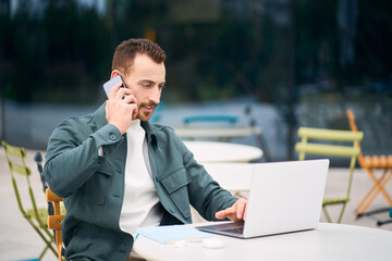 Confident handsome businessman talking mobile phone, using laptop and looking at screen sitting at workplace. Busy freelancer working online, typing 