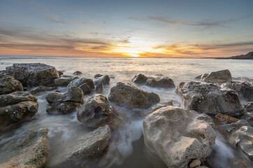 Ocean scene with low sun on the horizon, some rocks in foreground and mid ground with blurred waves around them plus a few colourful clouds.