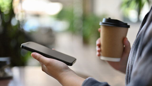 Cropped image of a female using her smartphone and sipping coffee while walking along the building.