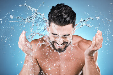 Water splash, hands and a man model washing his face in studio on a blue background for hygiene or...