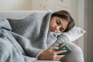 Moody sad teen girl wrapped in plaid lying on sofa at home, using smartphone. Upset teenage kid...