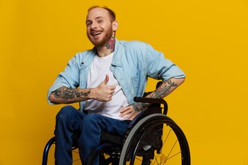 A man in a wheelchair smile and happiness, thumb up, with tattoos on his hands sits on a yellow studio background, the concept of health a person with disabilities