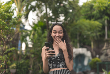 Fototapeta premium A pretty young Filipina woman with curly hair wearing a nice black top and long skirt giggling while chatting with a boy on her phone at the garden.