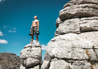 Strong muscular and fit white male standing on the peak of a mountain with kettlebells.