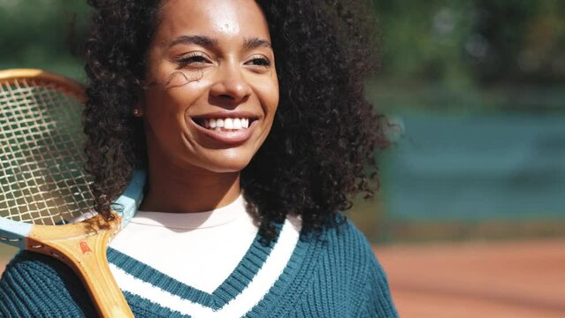 4k video. Young beautiful african american woman posing on clay tennis court with retro vintage tennis rocket. Sunny day outdoor. Sport, 80s, wellness concept.