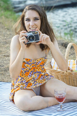 Happy smiling woman having picnic outside in nature wearing summer dress and photographs with vintage film camera - obrazy, fototapety, plakaty