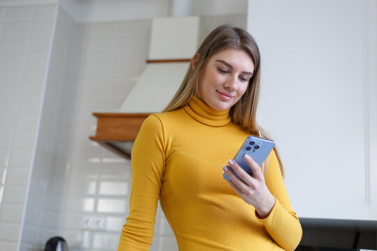 Happy Blonde Girl Using Modern Blue Smart Phone At Home. Portrait Of Beautiful White Woman In Early 20s Browsing Mobile App On A Cellphone