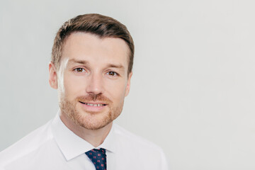 Portrait of attractive male CEO with confident facial expression, looks confidently directly at camera, wears elegant luxury clothes, poses against white studio wall with blank space on left for text