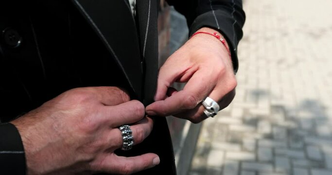 a man with a skull ring on his finger buttons up a black tuxedo jacket with a white stripe. close-up of male hands holding jacket