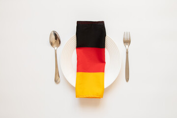 The flag of Germany lies on a white plate next to a spoon and fork on a white background, the...