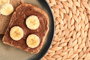 Plate of tasty toast with hazelnut butter and banana on wicker mat, closeup