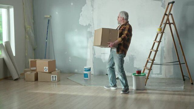 Elderly man is carrying heavy cardboard box, placing it on the floor and worried about back pain. Grandpa in plaid shirt is doing his own repairs and suffering from sciatica. Concept of repair in