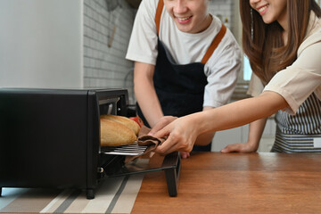 Cropped young couple taking off french baguette bread from oven, preparing dinner in modern kitchen together