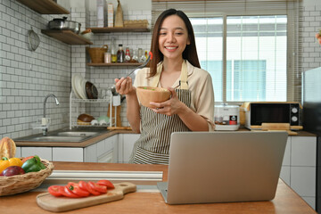 Attractive woman holding salad bowl and making video call on laptop in kitchen at home