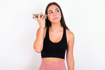 Smiling young woman wearing sportswear over white studio background listening a voice message from her smartphone. Communication and technology concept.