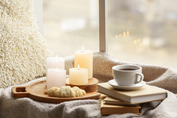 Fototapeta na wymiar Woollen cover with books, burning candles, croissant and cup of coffee at windowsill