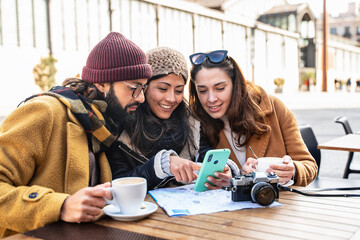 Young friends looking at cell phone sitting in cafe, checking tourist city map during winter...