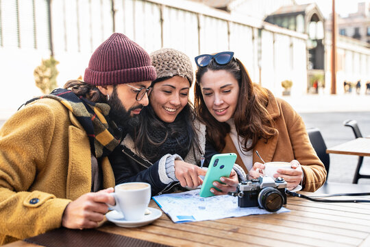 Young friends looking at cell phone sitting in cafe, checking tourist city map during winter vacations 
