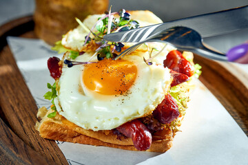 A woman cuts toast with scrambled eggs with yolk, sausages on a cutting board. Concrete background, morning light. Eggs for breakfast