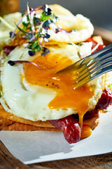 A woman cuts toast with scrambled eggs with yolk, sausages on a cutting board. Concrete background, morning light. Eggs for breakfast