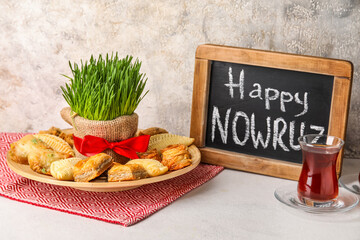 Chalkboard with text HAPPY NOWRUZ, treats, grass and glass of tea on table near grunge wall