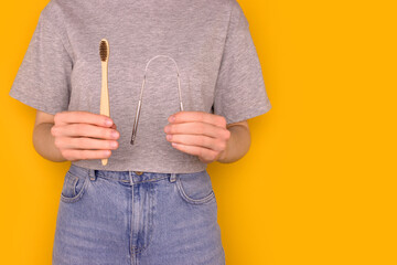 Female hands hold wooden toothbrush and metallic tongue scraper. Zero waste hygiene concept with copy space in front of yellow background. - obrazy, fototapety, plakaty