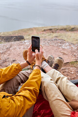 Close-up male hands holding smartphone taking photo of lake and nature surrounding, female hands on male legs, enjoy the rest together, travel concept