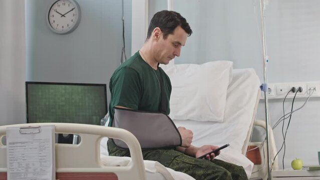 Caucasian soldier with arm bandage using smartphone while sitting on bed in hospital ward