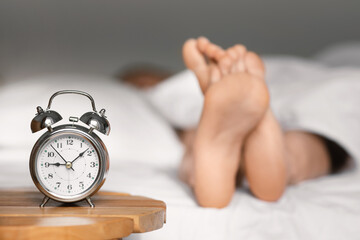 Feet of caucasian millennial lady lies on bed under white blanket, sleep at morning, focus on alarm...