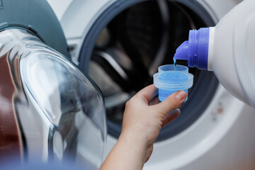 Woman pours liquid washing gel into plastic cap against background of drum of steel-colored washing...