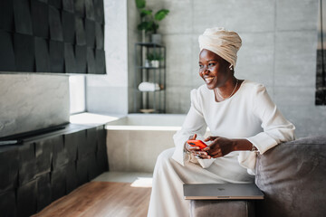 Happy African woman in white dress and turban holds phone sitting at home on couch. Happy African...