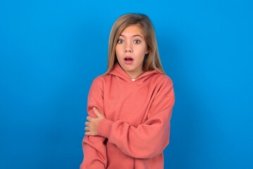 Fototapeta premium Shocked embarrassed caucasian teenager girl wearing pink sweater over blue background keeps mouth widely opened. Hears unbelievable novelty stares in stupor