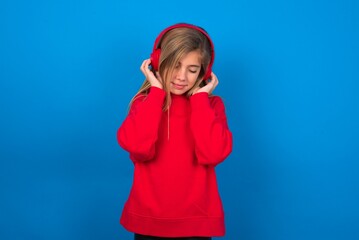 Pleased caucasian teenager girl wearing red sweater over blue background enjoys listening pleasant melody keeps hands on stereo headphones closes eyes. Spending free time with music