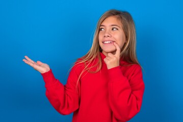 Positive caucasian teenager girl wearing red sweater over blue background advert promo touch finger teeth