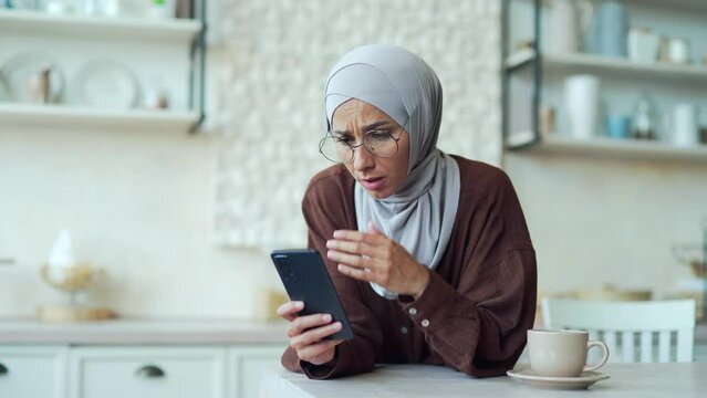 Scared young muslim woman in hijab read bad news browsing social media in smartphone while sitting at home Upset Confused female stares at screen and looks puzzled, unexpected problem fear
