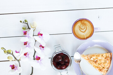 cup of  cappuccino coffee, pie on the violet plate, silver tea spoon, orchid flower, jar with strawberry jam on white colored wooden table,  top view - 569162871