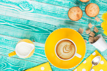 cup of cappuccino coffee on yellow plate and yellow milk jug cane sugar,  cakes, dandelions, teaspoon on turquoise colored wooden table with yellow napkin at polka dots top view - 569162887
