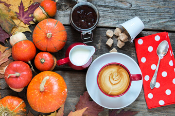 cup of cappuccino coffee milk jug, strawberry jam, red napkin at polka dots on wooden table decorated by falling leaves and pumpkins, top view - 569162896