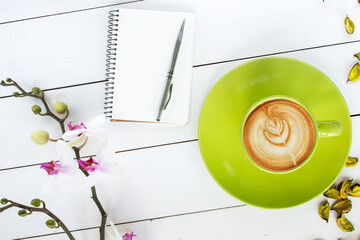 cappuccino coffee in green cup,  orchid flower, notebook and silver pen,  green dry flower decor scattered on white painted wooden table, top view - 569162897