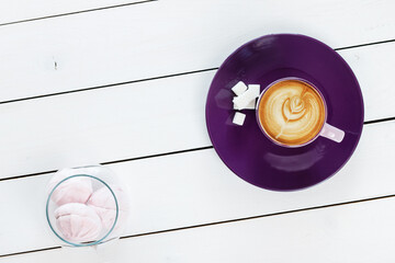 cup of cappuccino coffee on violet plate with sugar,  marshmallows in a glass vase on white colored wooden table , top view - 569162899