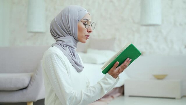 A young Muslim woman in a hijab prays at home. Alone in a bright living room in the house worshiping to the GOD reading Quran while wearing veil in the lounge Eastern culture religion lifestyle