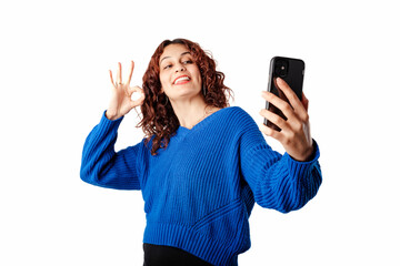 Happy woman wearing sweater standing isolated over white background taking a selfie with the okay sign, looks at the phone and smiles. Video call with family. Video call with hearing impaired friend.