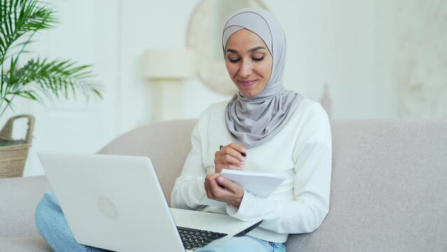 Smiling young muslim woman in hijab student having a remote lesson listening teacher lecturer making notes video course and looking at laptop screen in living room indoor Distance online education