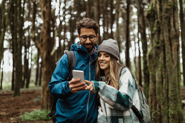 Smiling young european guy and woman tourists in jackets with backpack walk in forest, make route