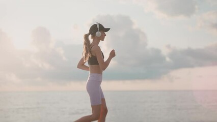 A young woman runner is listening to music in earphones and training on a sea beach. Close up of fitness girl is jogging outdoor. Concept of workouts running and healthy lifestyle. Slow motion