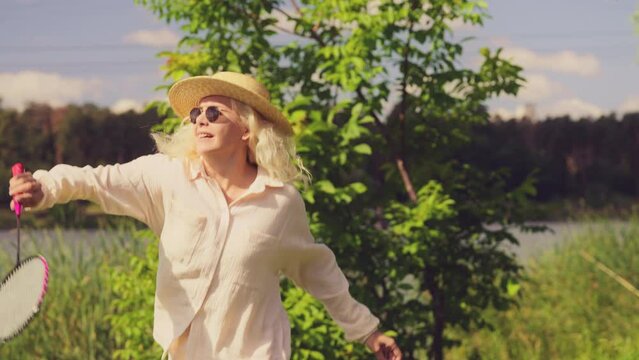 Smiling middle-aged blond woman playing badminton by the lake, outdoor activity